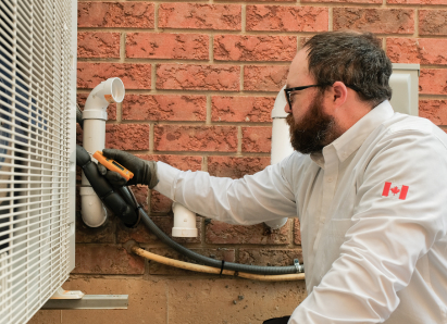 A Francis HVAC specialist wearing glasses and gloves uses a diagnostic tool to service an outdoor AC unit beside a brick wall.